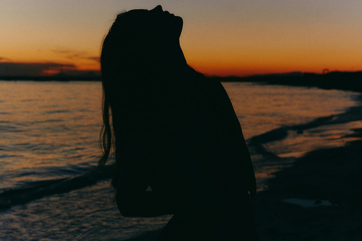 A person on a beach, silhouetted against the sunset, throwing their hair back and looking up towards the sky. Illustrating an interview with Sarah Wilson about her book, This One Wild and Precious Life.